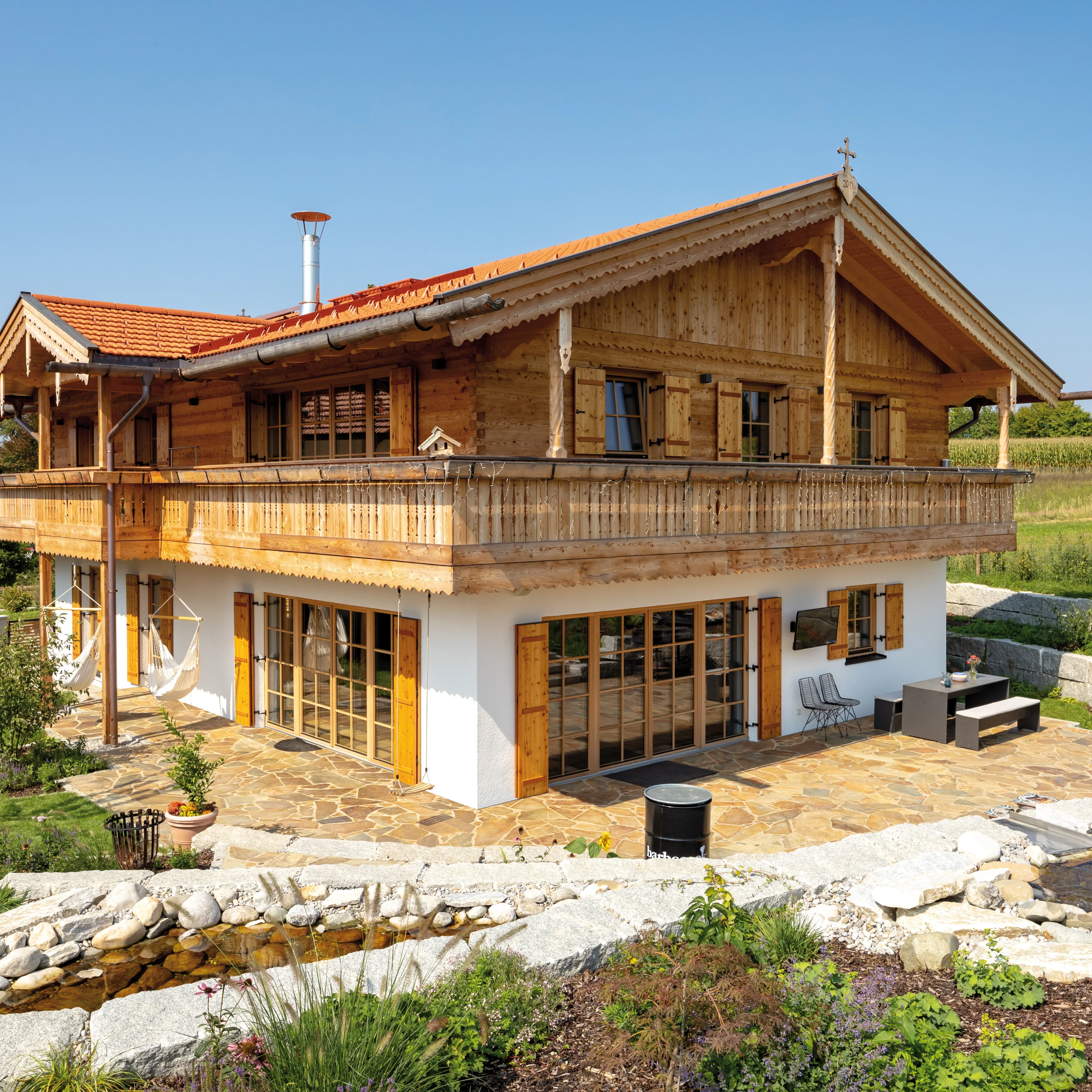 Ein traditionelles Holzhaus mit weißem Putz und einem roten Ziegeldach, Balkon, Terrasse und Garten mit Bachlauf.