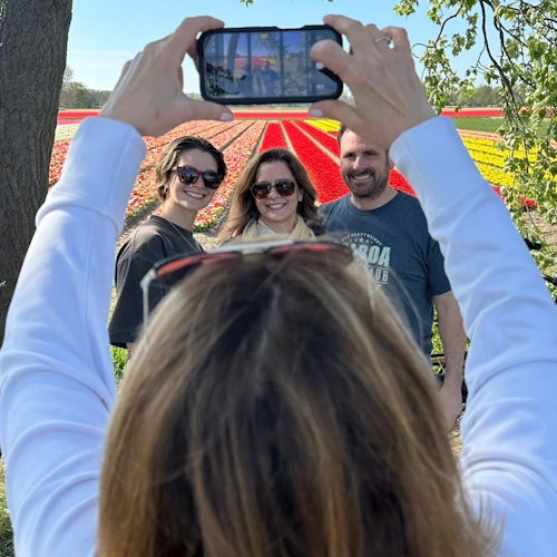 Kuhang larawan ng isang tao kasama ang tatlong nakangiting indibidwal na nakatayo sa harap ng makukulay na palayan ng bulaklak. May puno at mga sanga sa likod.