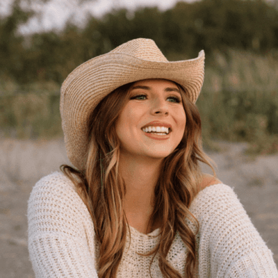 Portrait of Ashlie Discenza smiling outdoors, wearing a straw hat and a light knit sweater with a natural landscape in the background.