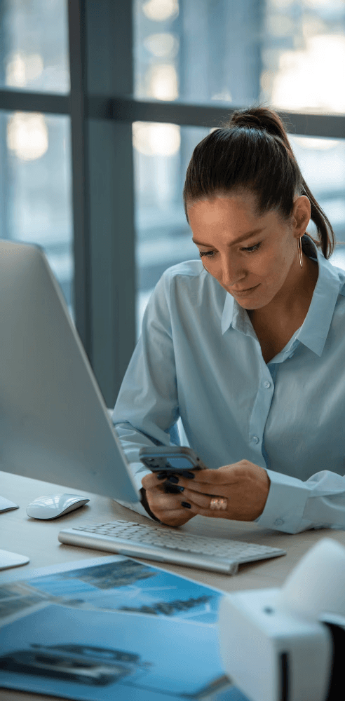 Woman working at a desk with a smartphone and laptop, representing efficient project planning and execution.