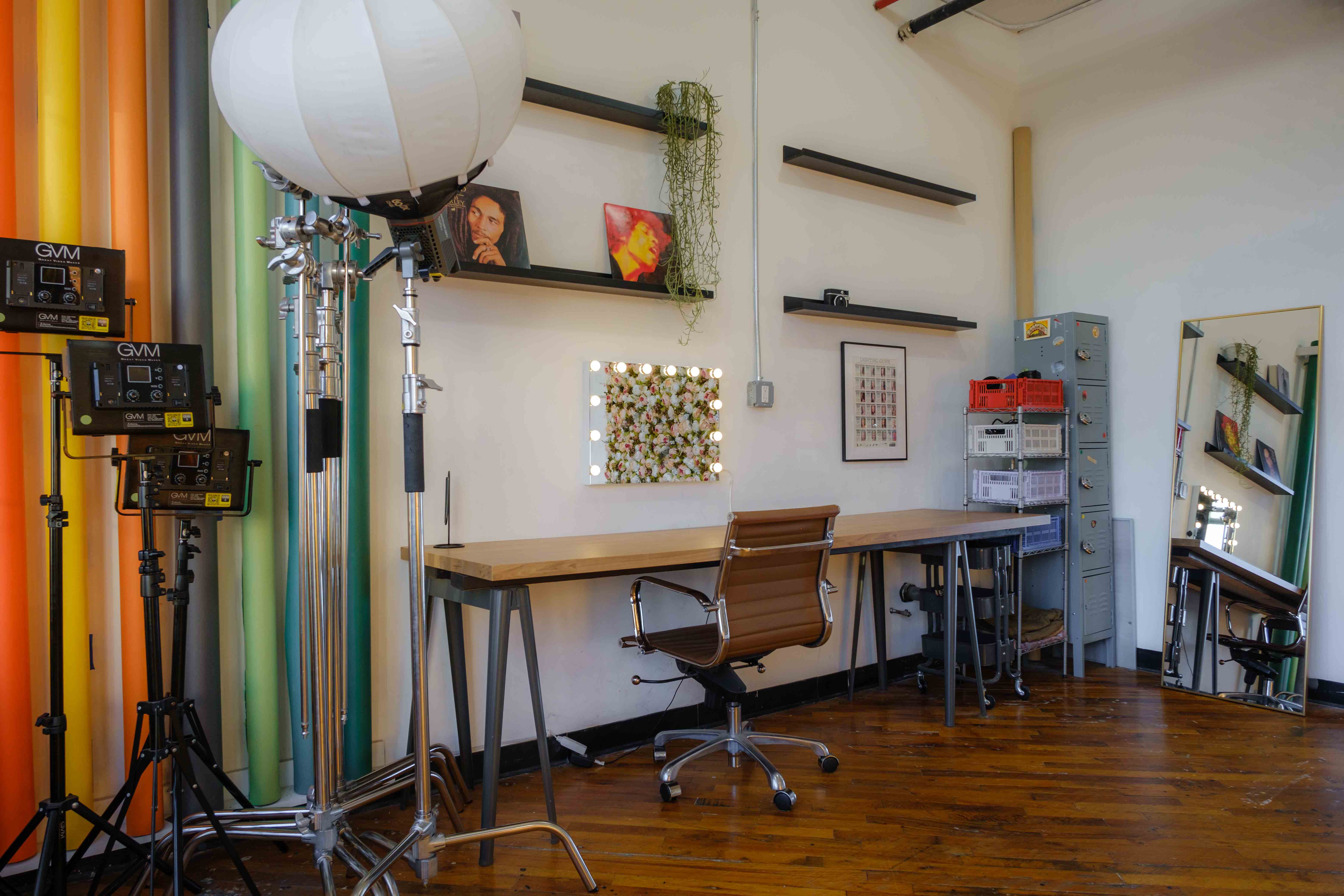 A workspace in a daylight photography studio, featuring a long wooden desk, an ergonomic chair, and lighting equipment set against colorful backdrops. The area includes shelves with decor, a floral-framed mirror with bulbs, and a storage unit, combining functionality with creative flair.