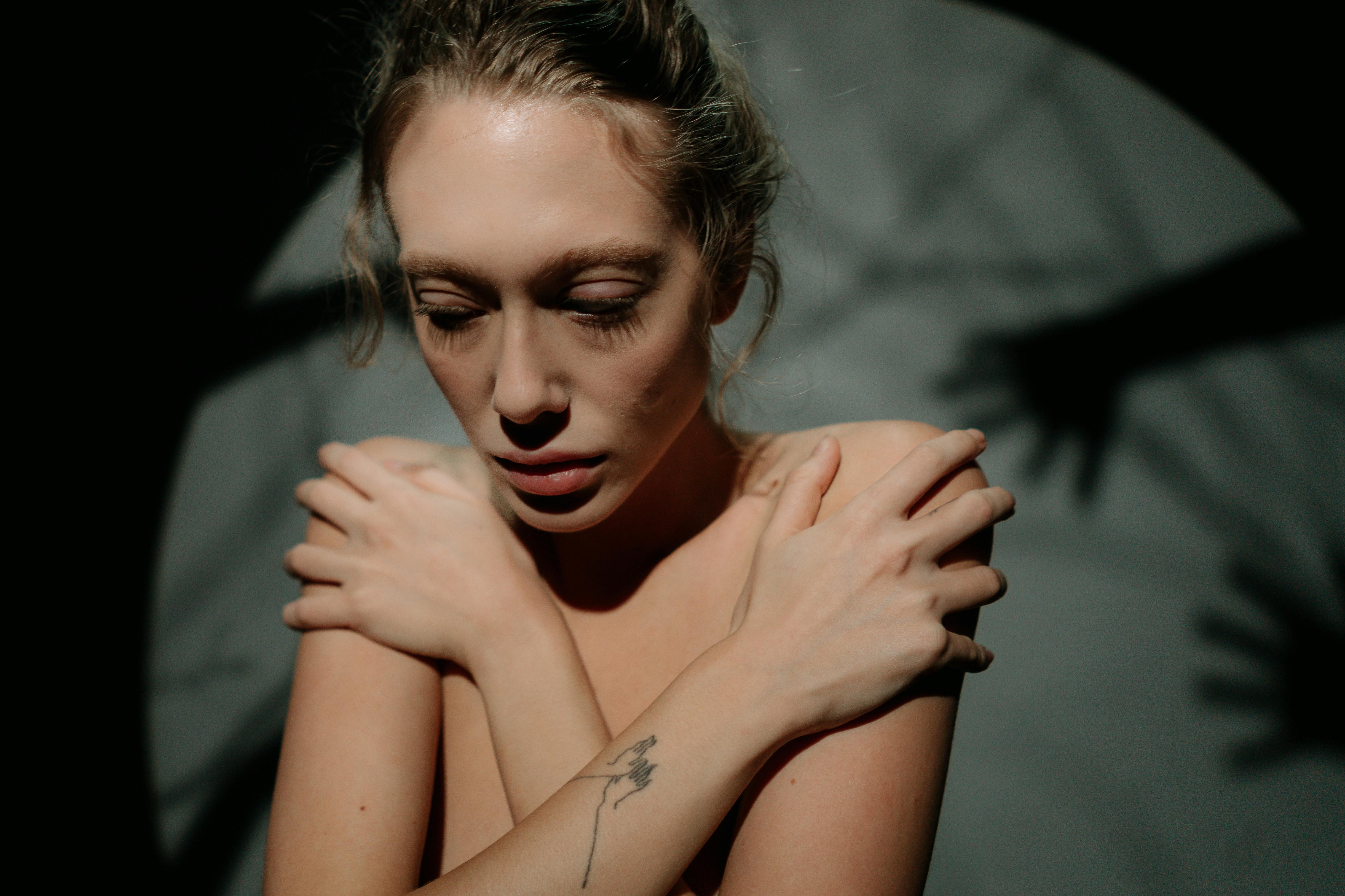 A woman looking very tense and uncomfortable in her body. She is covering her breast and grabbing her shoulders with her hands while looking down as she is scared of being judged. It is represented by shadows of hands all around her in the background