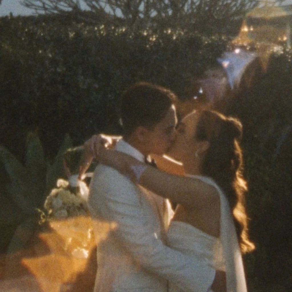 A couple shares a romantic kiss in a Super 8 Film still during their wedding ceremony, surrounded by greenery and bathed in dreamy, golden sunlight, with a bouquet of white flowers adding a touch of elegance.