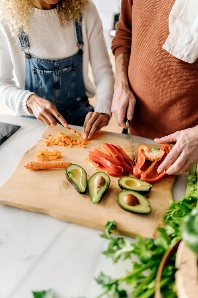 Two people are preparing a fresh, colorful meal with sliced red bell peppers, halved avocados, and a chopped carrot on a wooden cutting board, surrounded by leafy green vegetables.