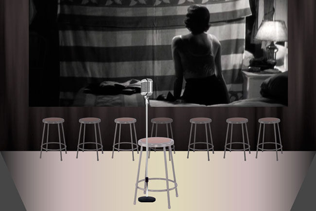 A black and white image shows a woman sitting with her back to the camera, facing a large screen displaying a striped curtain. A microphone on a stand is positioned in front of her. Six light-colored wooden stools are arranged in front of the screen, and a lamp is visible on a small table to the right. The floor is light-colored, and the image is rendered in grayscale.