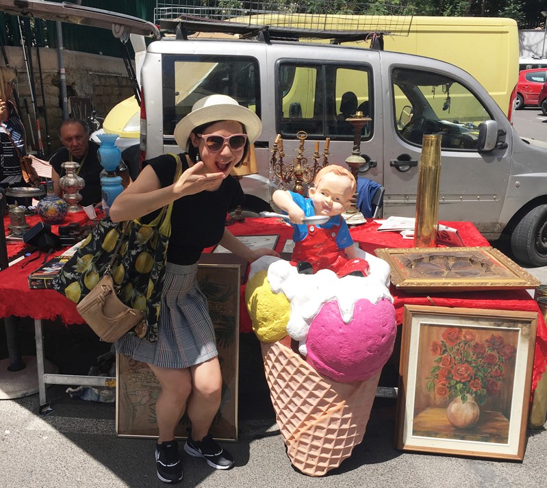 A photo of a woman sitting in front of an artisan stand near a giant ice-cream cone with a baby on top that has a spoon.