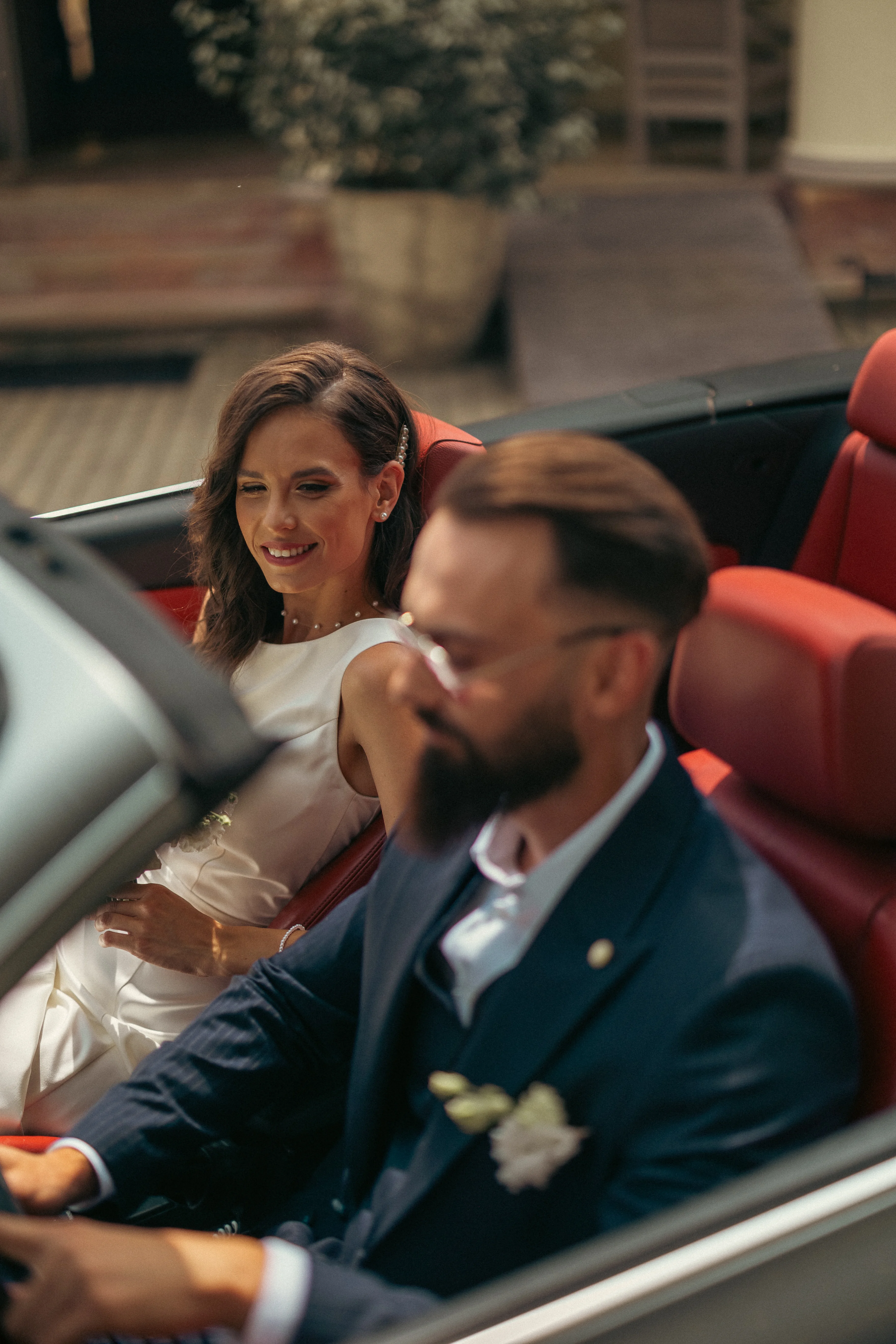 Candid esküvői fotó: mosolygó menyasszony fehér elegáns ruhában és vőlegény kék öltönyben ülnek vintage kabrióban piros belső téréssel, Fenyőharaszti Kastélyszálló