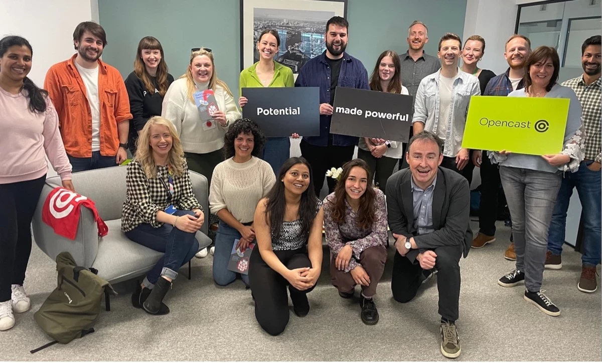 A large group of people gather in an office space, some holding signs that read “Potential made powerful” and “Opencast.” Others stand or sit at the front, with a sofa, backpacks, and a framed city photo visible in the background.
