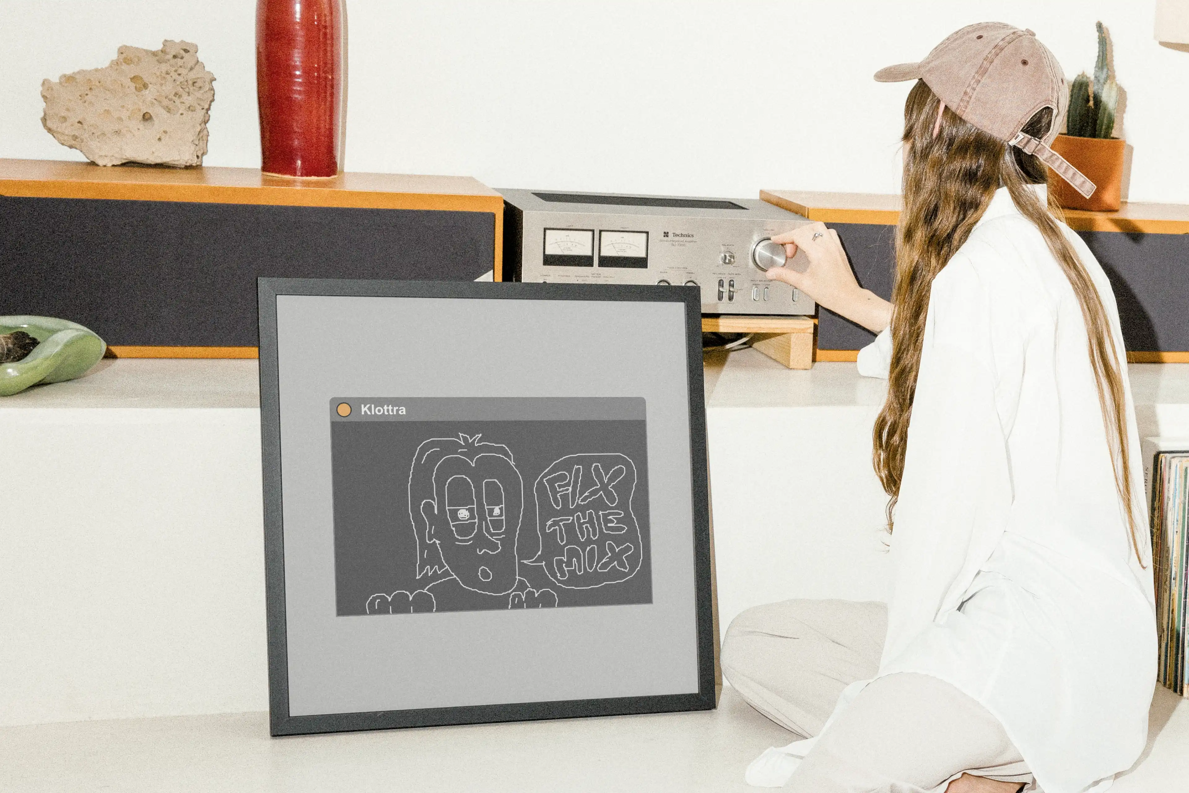 Framed Klottra doodle print displayed on the floor beside a woman adjusting a vintage audio receiver.
