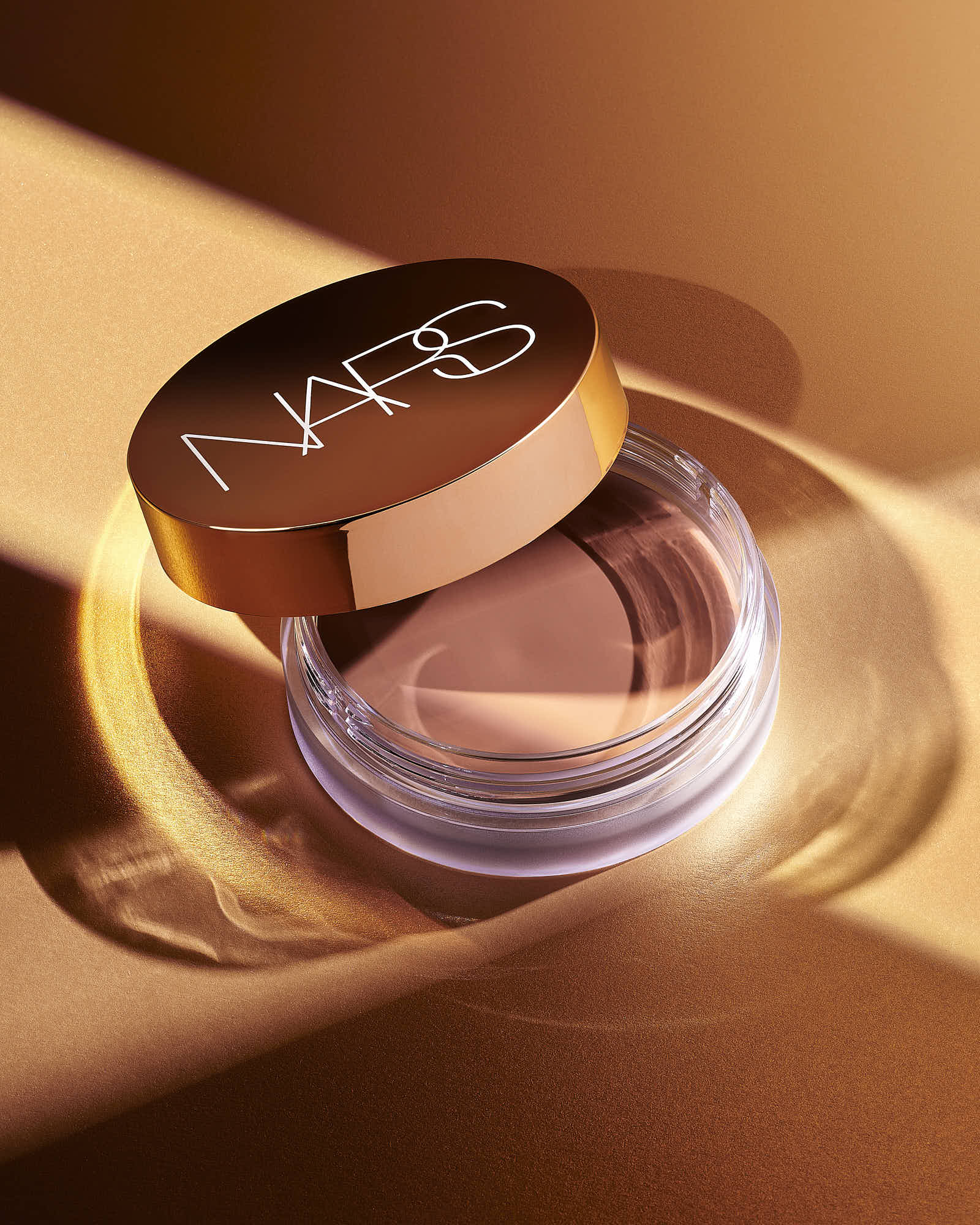 A makeup product with a bronze lid opened to reveal a powder inside, set against a warm golden background.