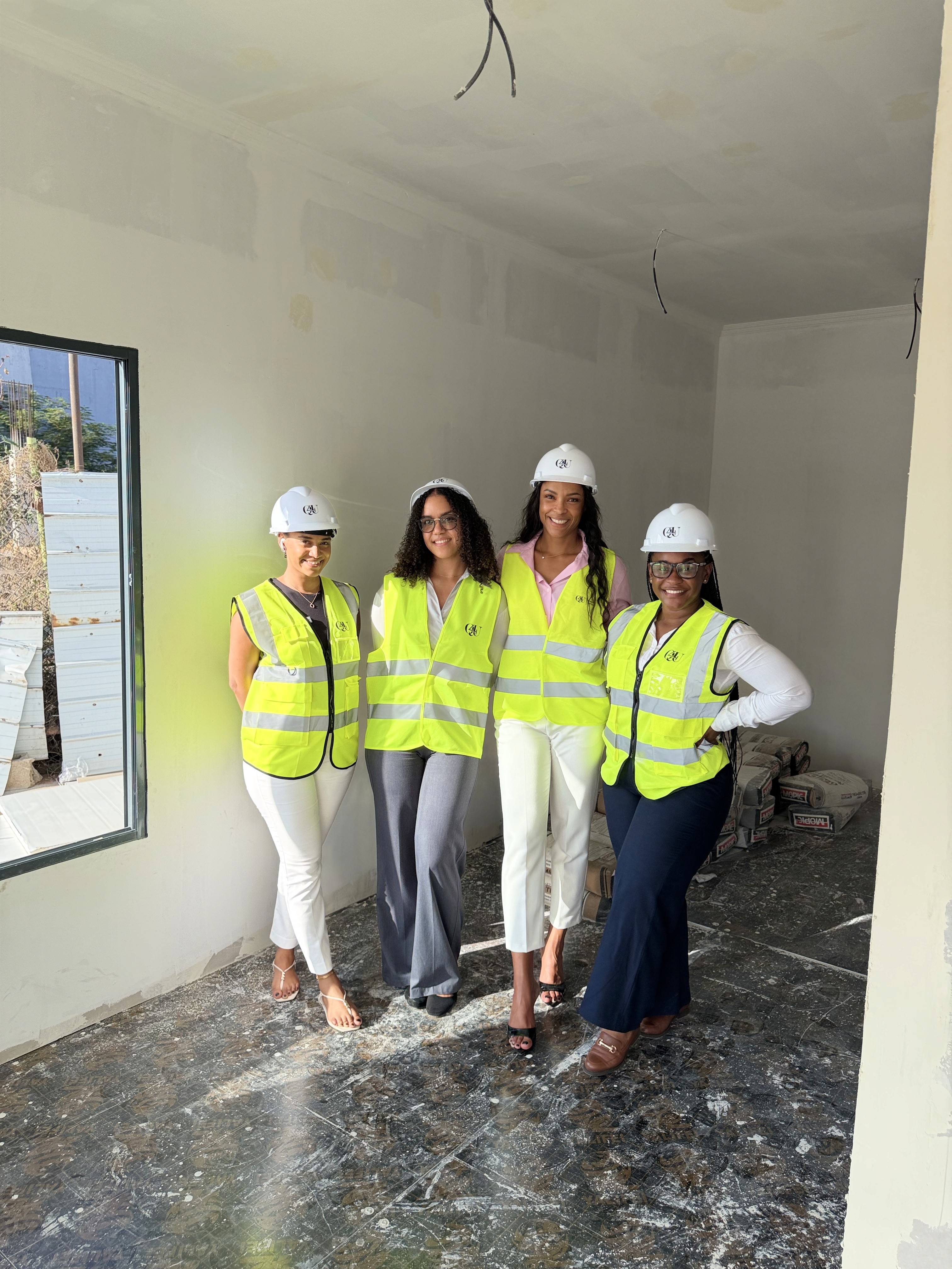 Group of four construction professionals in safety vests and helmets smiling and shaking hands on a worksite, conveying teamwork and collaboration.
