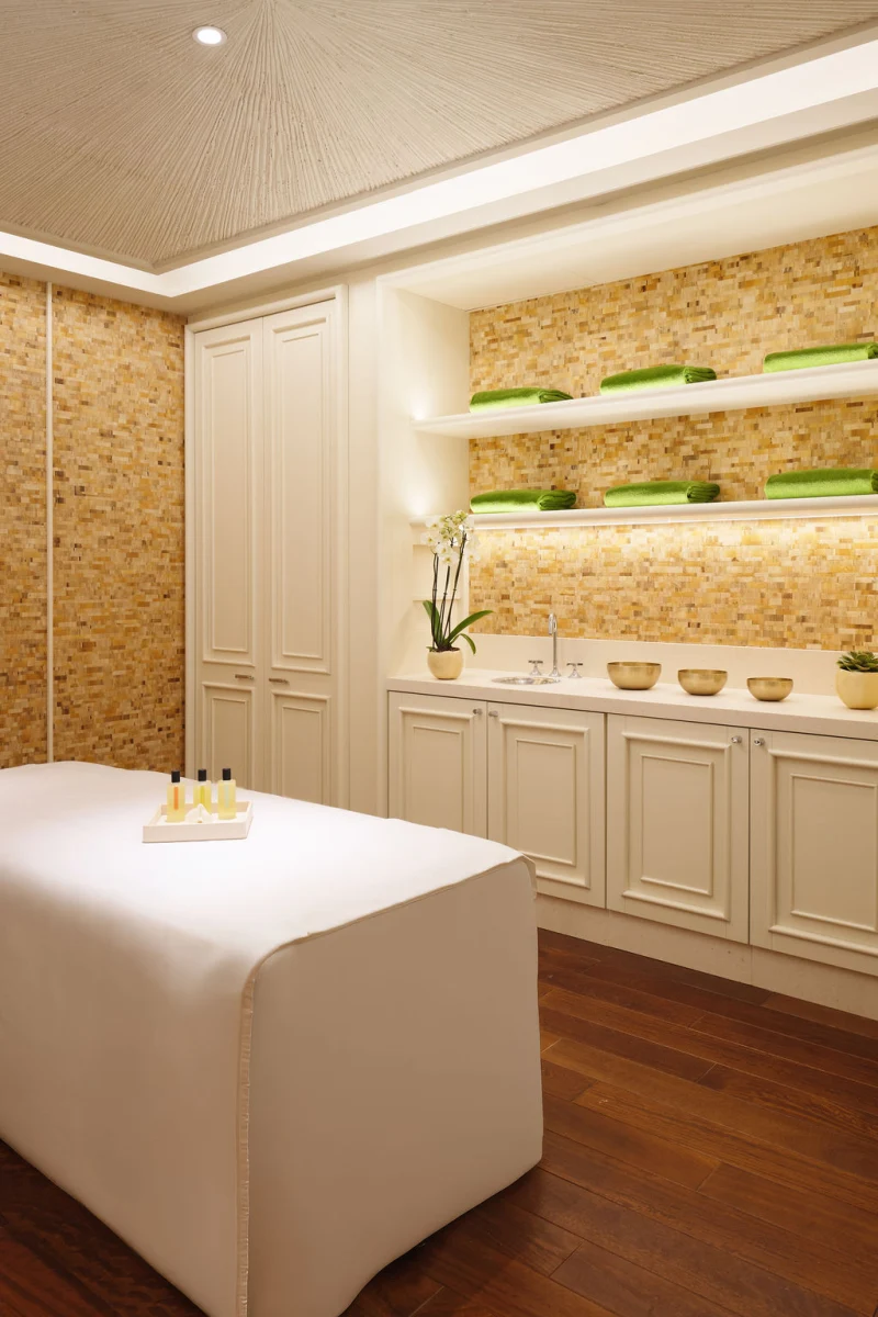Ein ruhiger Behandlungsraum im Spa mit einer Massagebank, warmen Holzböden und beleuchteten Regalen mit grünen Handtüchern und goldenen Schalen.