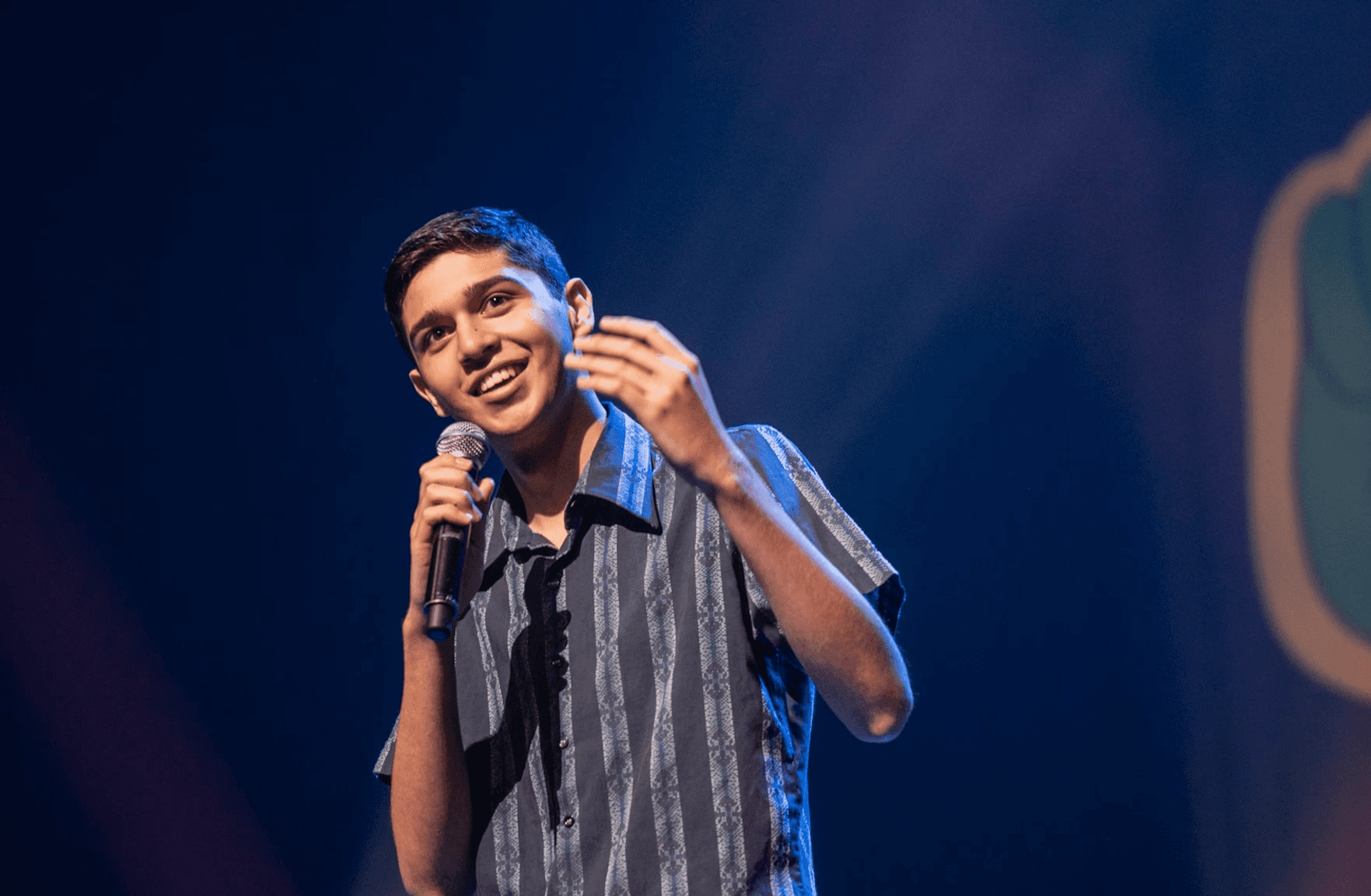 A young person stands onstage holding a microphone, smiling as they speak to an audience. They wear a short-sleeved patterned shirt, lit by colourful stage lighting. A large graphic of a cartoon hand making a peace sign is visible in the background, suggesting a lively performance or presentation event.