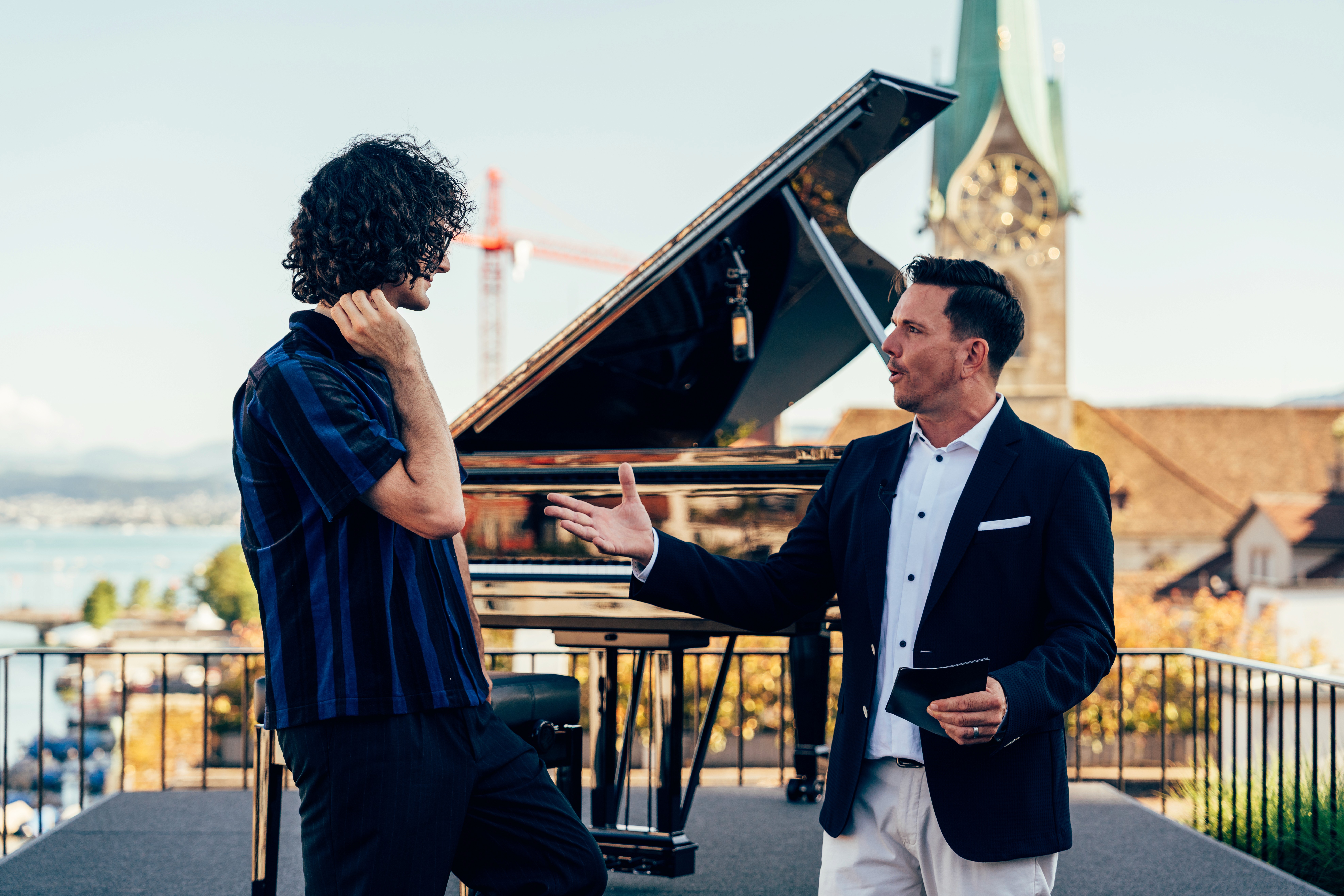 Das Foto zeigt zwei Männer im Gespräch auf einer Dachterrasse oder erhöhten Plattform an einem strahlend sonnigen Tag. Der Mann auf der linken Seite hat dunkle Locken, trägt eine Brille und ein dunkelblau-schwarz gestreiftes Kurzarmhemd. Er fasst sich mit einer Hand leicht an den Hals oder Nacken. Ihm gegenüber steht ein Mann in einem dunklen Sakko über einem weißen Hemd. Dieser gestikuliert offen mit der rechten Hand und hält in der linken Hand kleine dunkle Notizkarten, was stark auf eine Interview- oder Moderationssituation hindeutet. Genau zwischen und hinter den beiden steht ein glänzender schwarzer Flügel. Im Hintergrund erstreckt sich eine malerische Kulisse mit einem See, fernen Bergen und Stadtarchitektur, aus der ein markanter, spitzer Kirchturm mit Zifferblatt (vermutlich das Fraumünster in Zürich) in den blauen Himmel ragt.