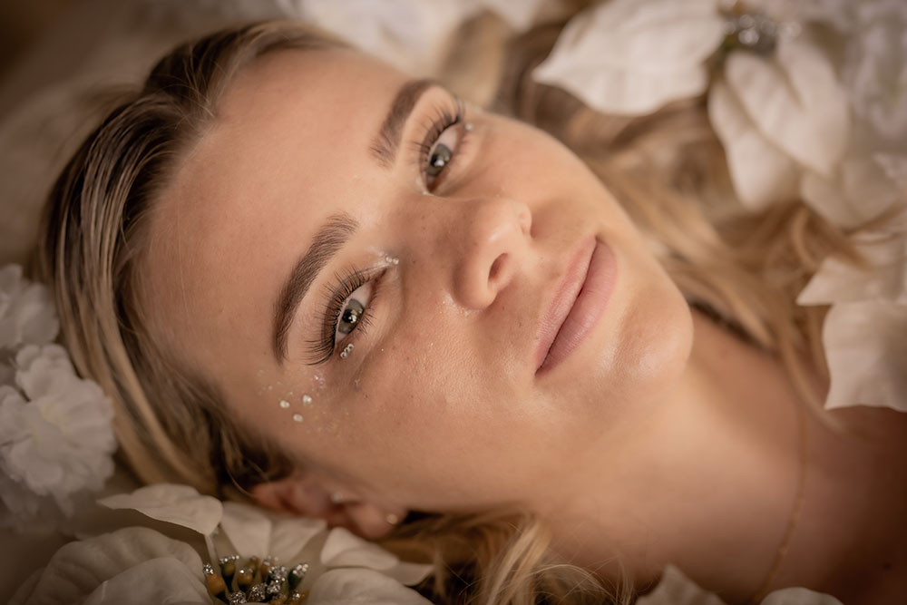 Close-up portrait of a person wearing a gray insulated hooded jacket with the hood up, showing their face framed by the hood against a light background.