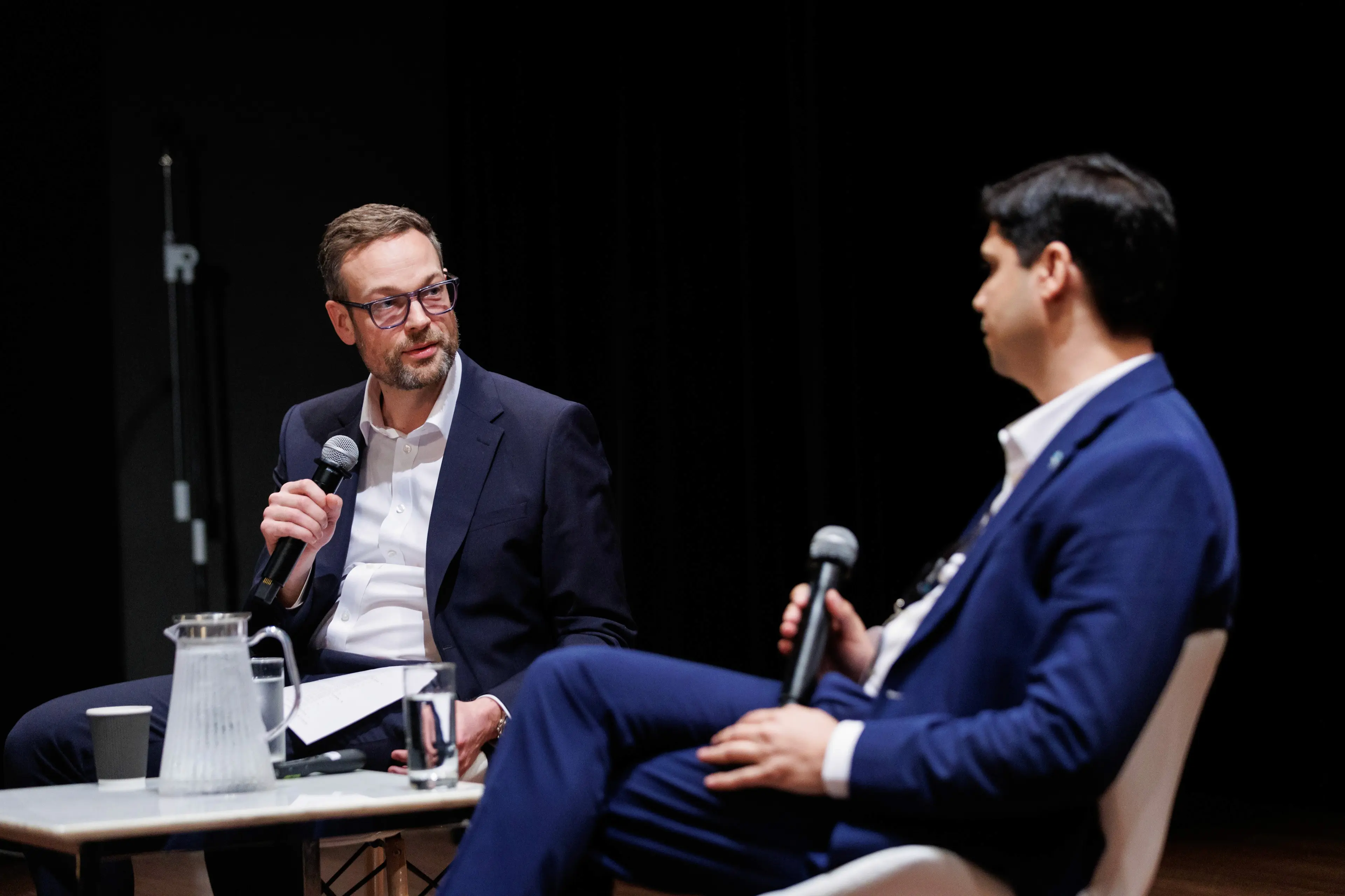 Two men seated on stage engaged in a discussion, one holding a microphone and the other listening attentively.
