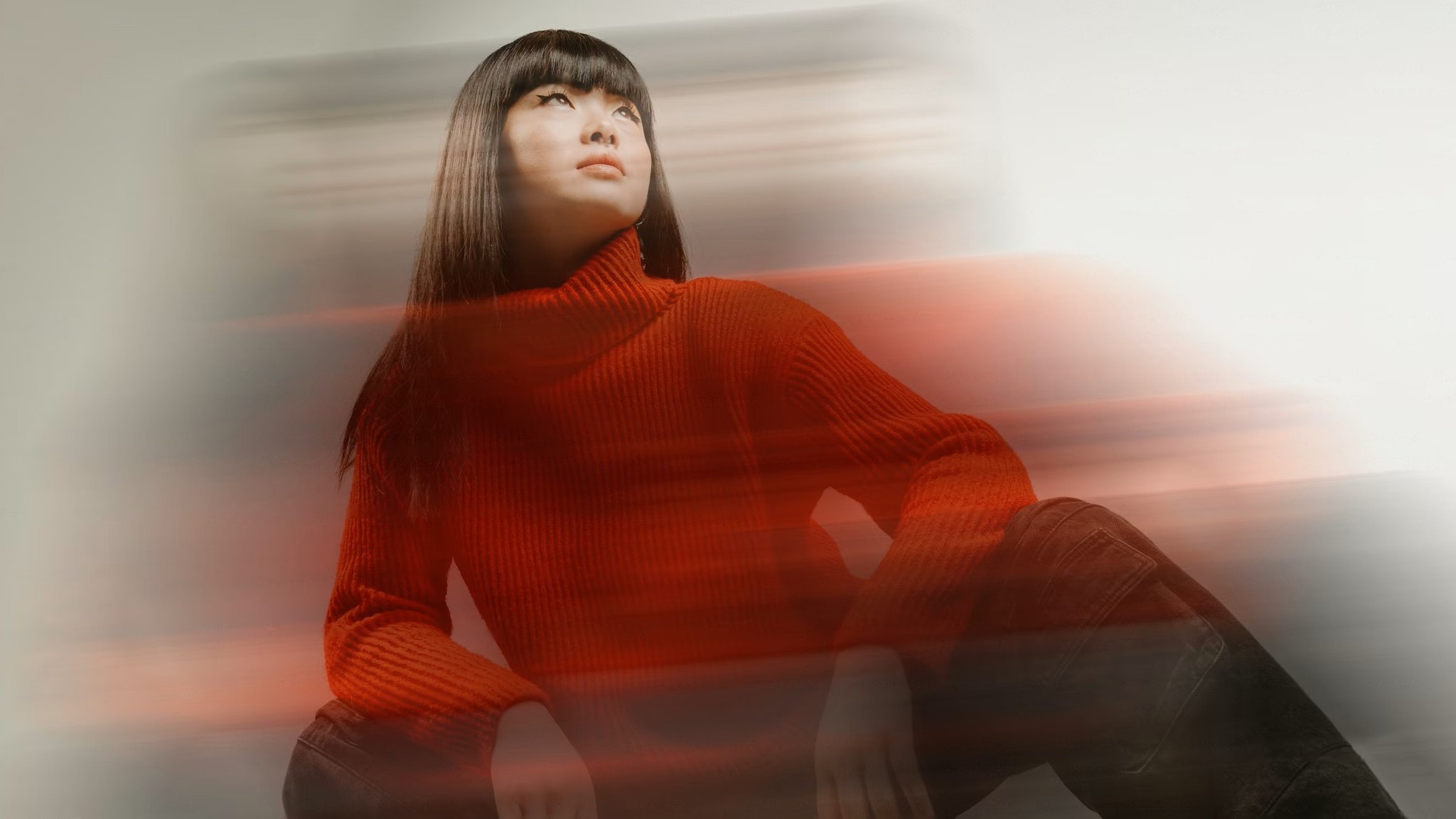 A person wearing a vibrant red sweater is seated against a blurred, dynamic background, creating a sense of motion and energy.