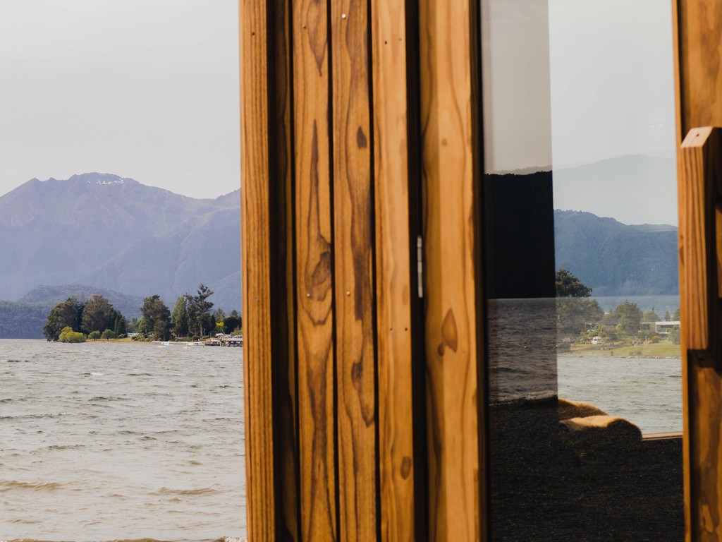 View through a wooden window shows a serene lake with gentle ripples and distant mountains shrouded in mist, creating a peaceful and inviting scene.