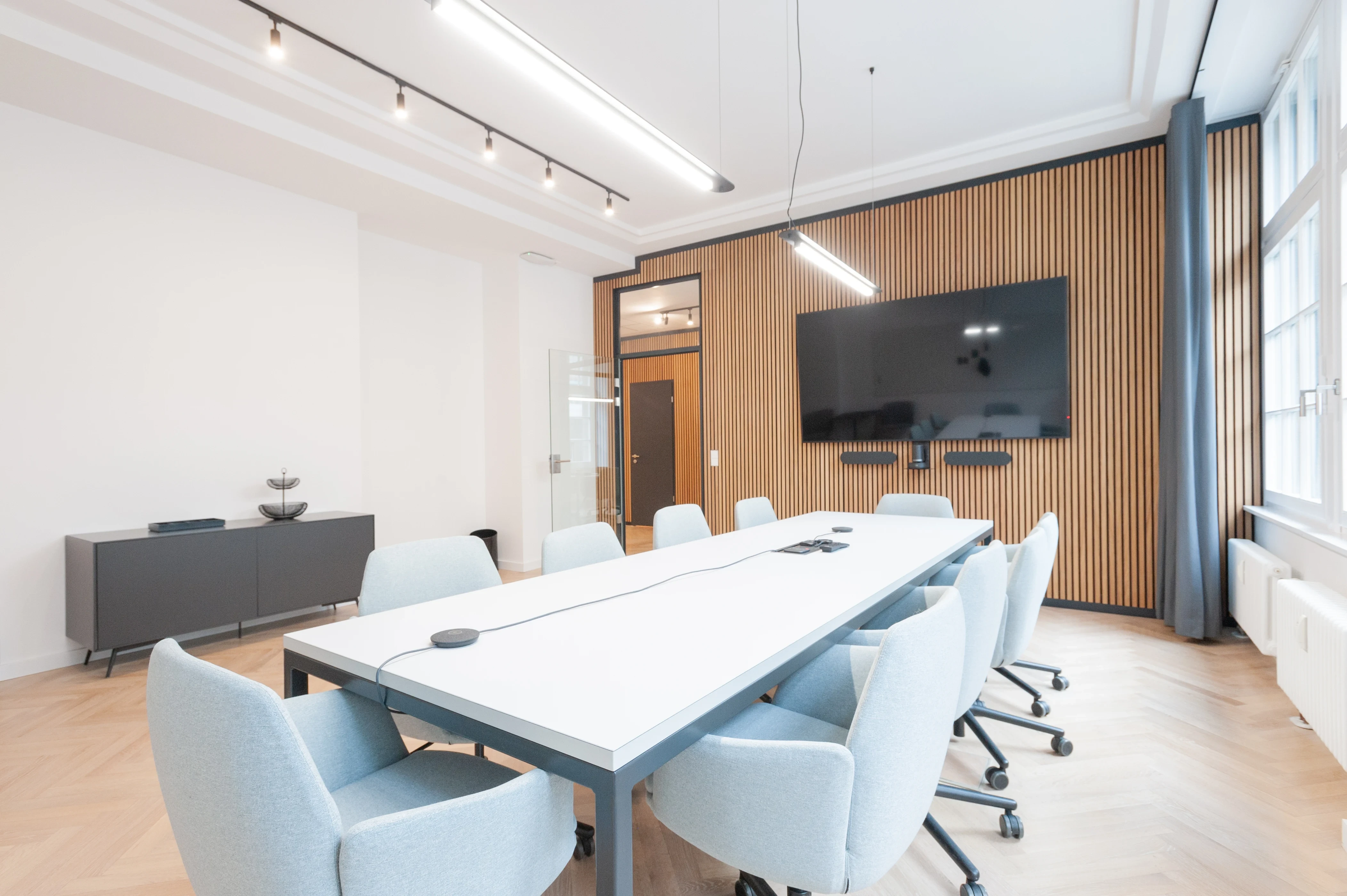 Conference room at AI START-UP BERLIN with light blue chairs around a long white table with cable hatches, a TV on an oak slat wall, a grey sideboard and high stucco ceilings.