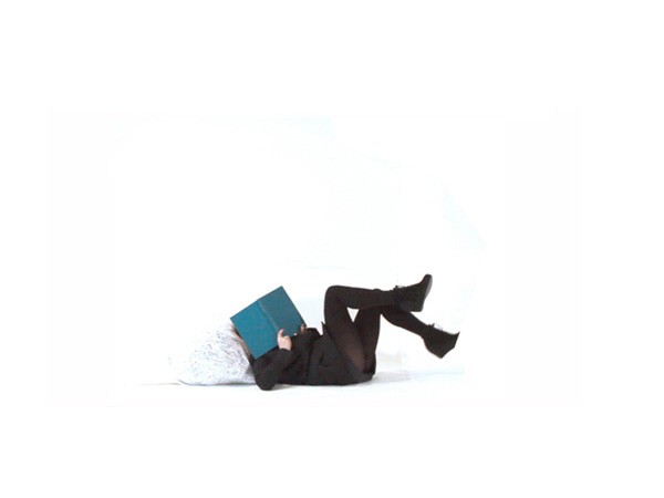 A person lies on their back on a white surface, wearing a black long-sleeved top, black pants, and black ankle boots. Their legs are bent at the knees and raised slightly. A teal-colored book is held open in front of their face, obscuring their features. The background is entirely white.