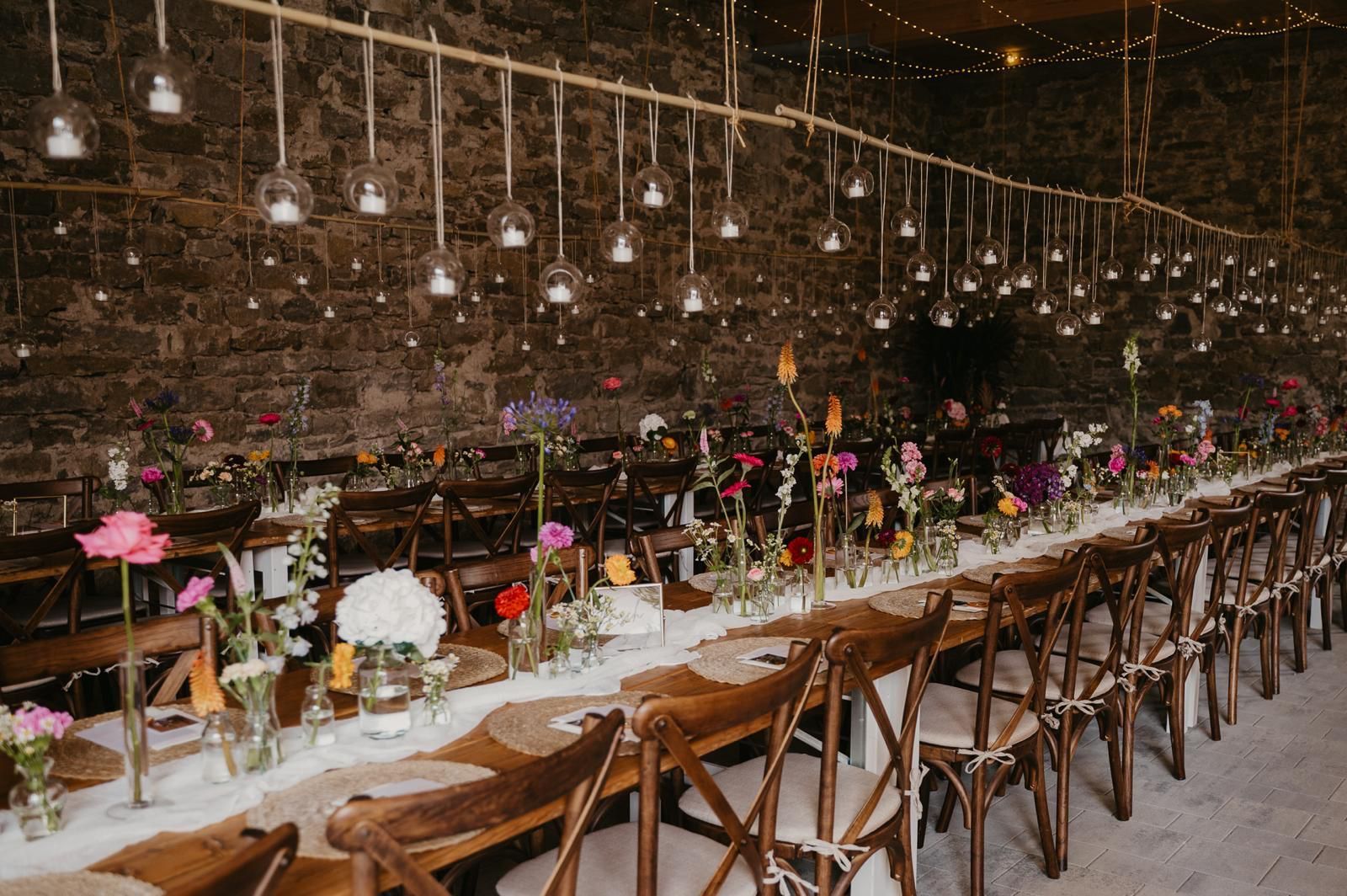 Lange Holztafel vor einer Natursteinwand mit hängender Glühbirnen-Beleuchtung und Vintage-Mietmöbeln