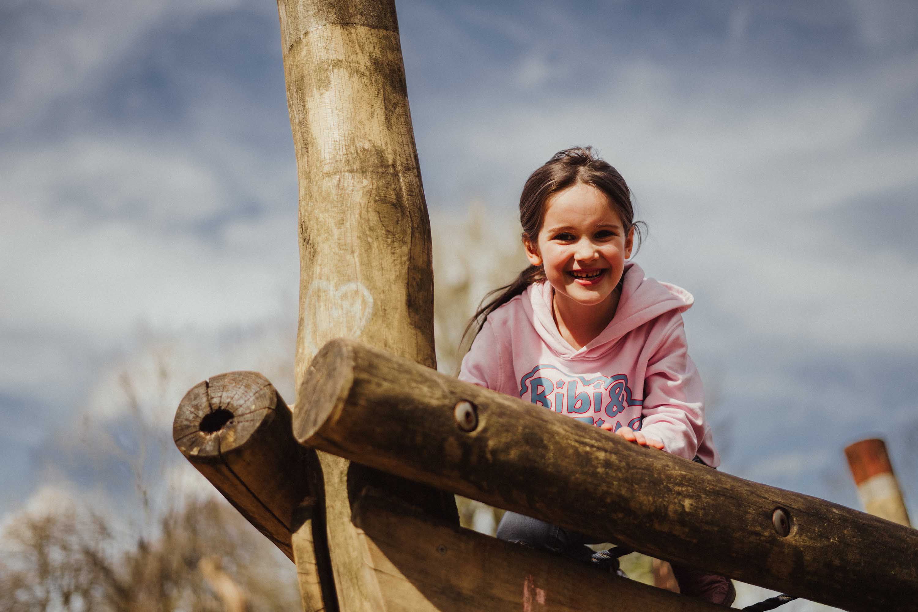 Authentische Kindergartenfotografie Gevelsberg: Ein Mädchen in einem rosafarbenen Pullover lacht glücklich beim Klettern auf einem Holzgerüst. Professionelles Outdoor-Porträt mit weichem Bokeh von PK photography 4 Kids NRW.