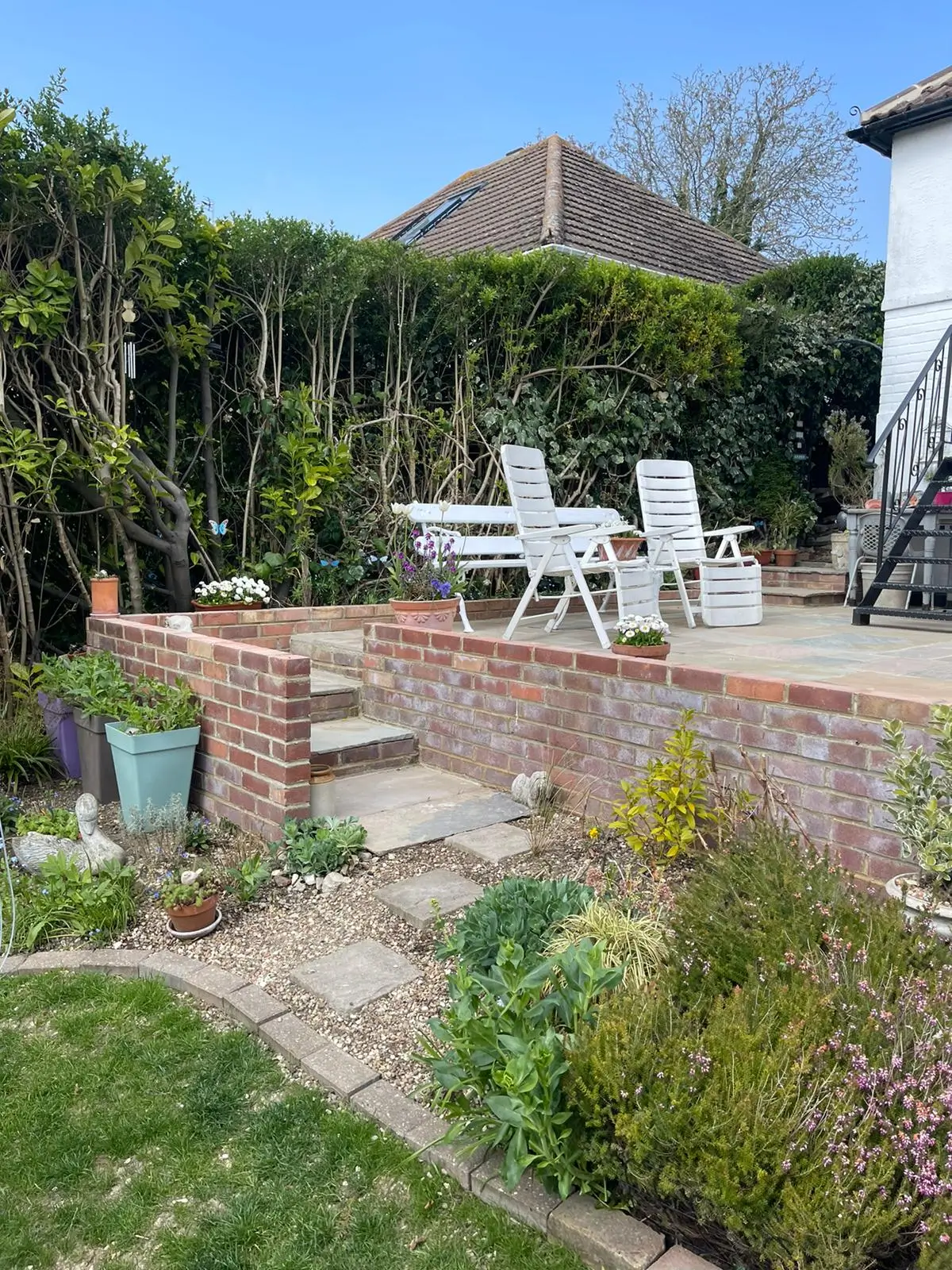 A sunny garden scene featuring a pathway, greenery, and seating area surrounded by hedges and foliage.