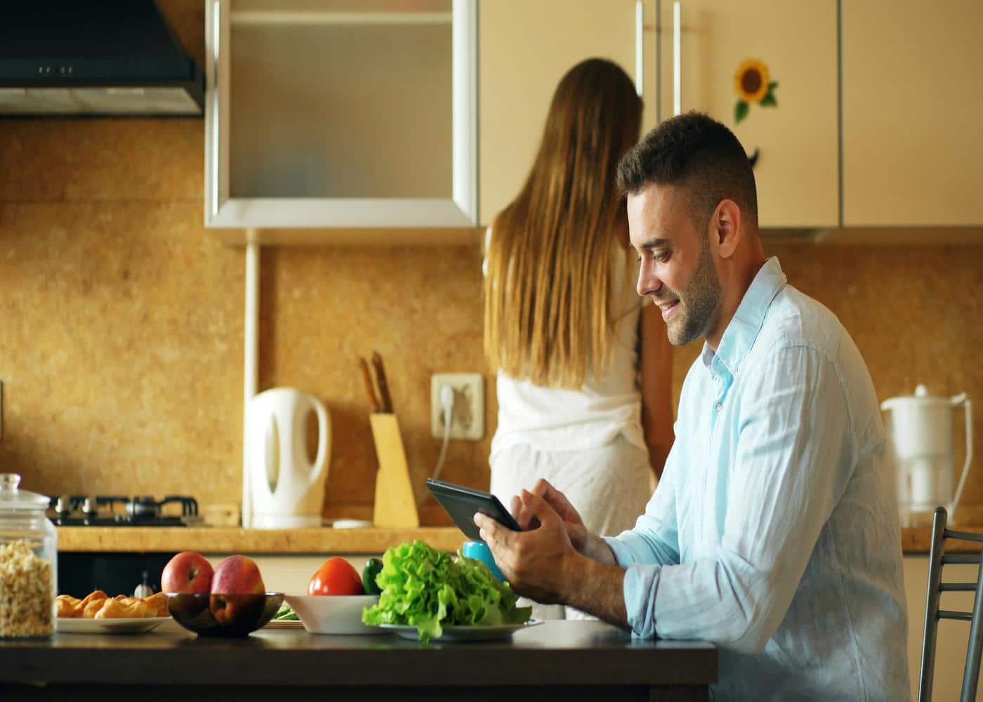 Man using tablet in kitchen with nutritoious food on the table and woman behind him