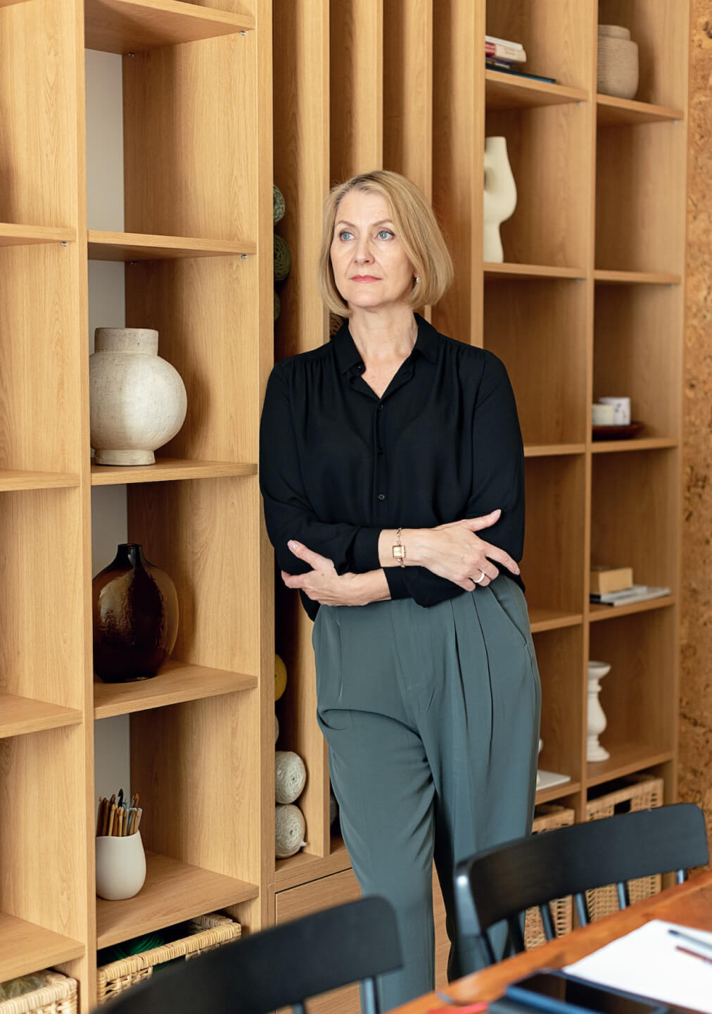A woman in a black blouse and grey trousers stands confidently with arms crossed, in front of wooden shelves with decorative pottery in a modern office.