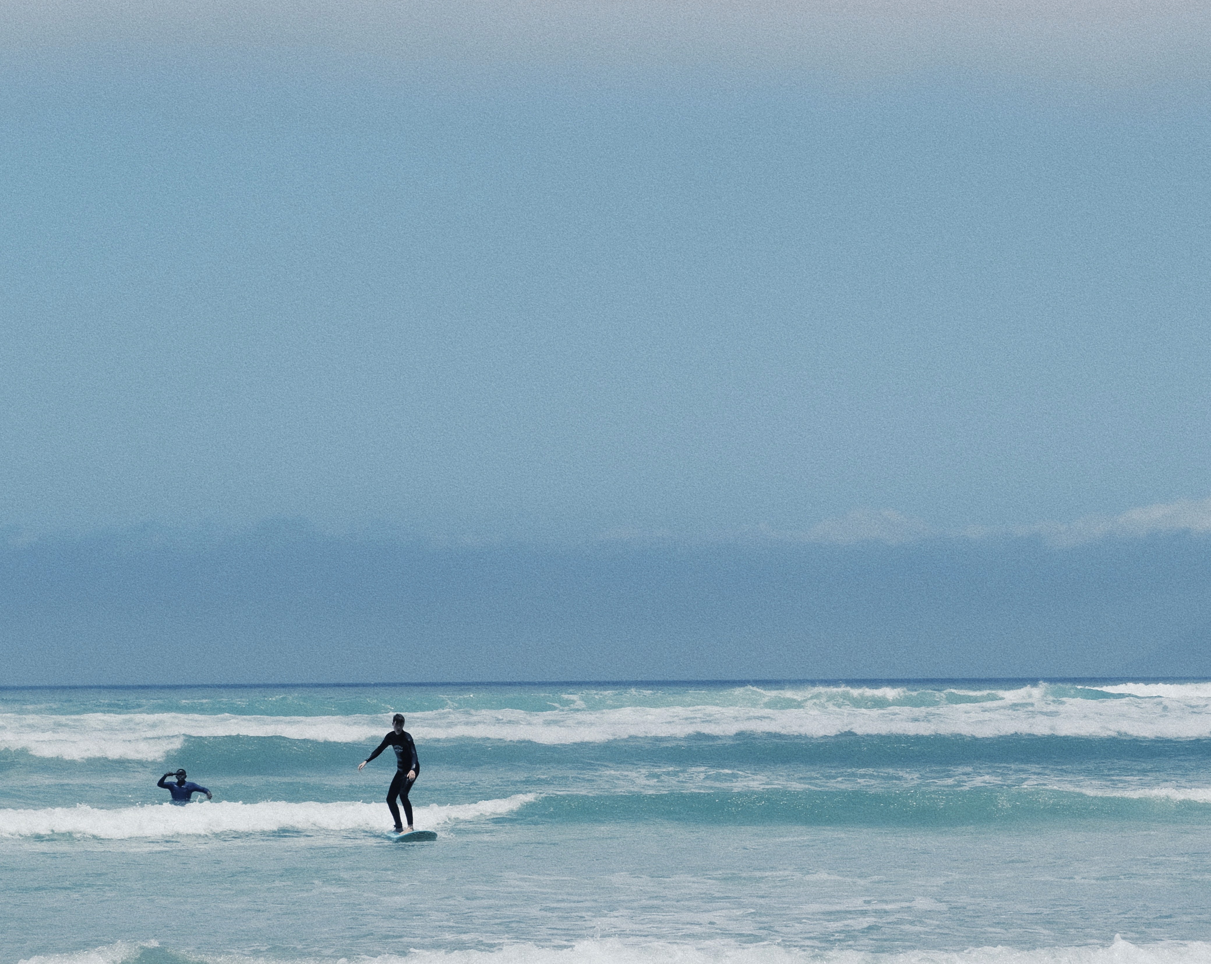 A surf student surfing a wave while his coach is behind him, filming him, during their surf lesson in Muizenberg, Cape Town