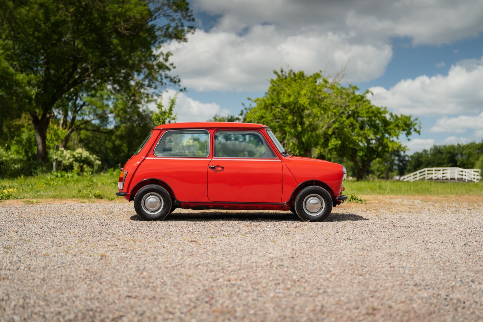 A bright red vintage car parked on a gravel road, surrounded by green trees and a cloudy sky.