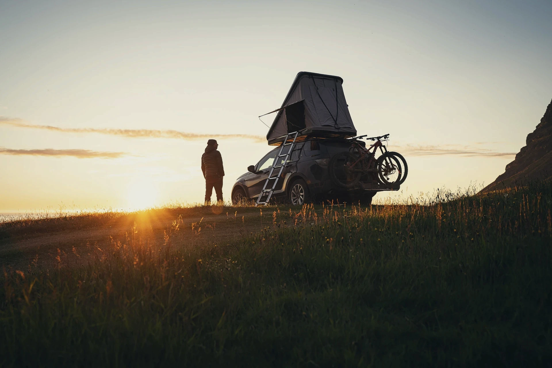 Car with rooftop tent and bikes parked at sunset, person standing nearby.