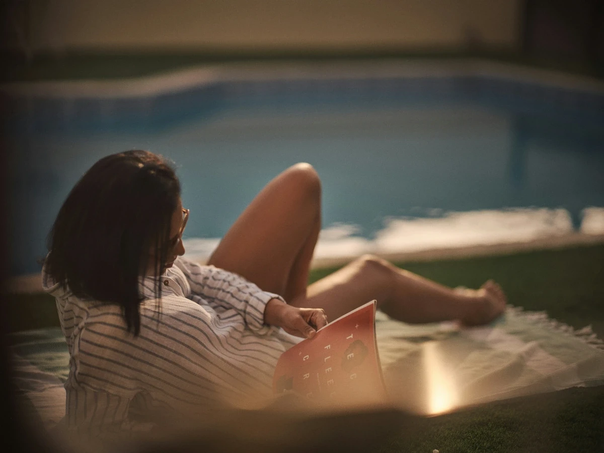 Mujer joven relajándose y leyendo un libro sobre una manta en el césped junto al borde de una piscina iluminada de noche.