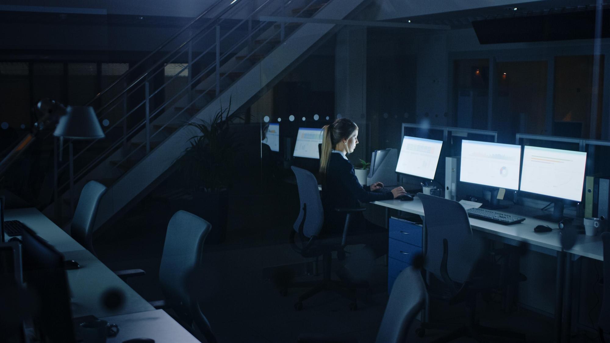 A woman works at her desk is a dark, empty office