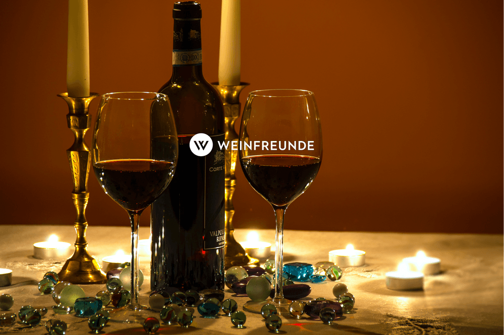 Ein Tisch in einer gemütlichen Atmosphäre der dekoriert ist mit Teelichtern und bunten steinen sowie zwei Kerzenständern, darauf ist eine Weinflasche mit zwei Weingläsern gefüllt mit Rotwein, darauf befindet sich das Logo der Weinfreunde