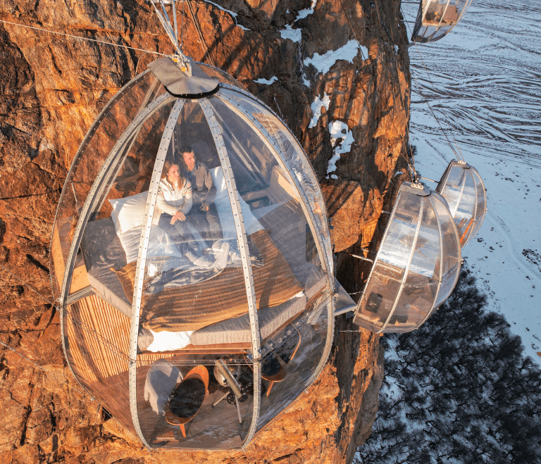 Pareja sentada en cama dentro de cápsula transparente colgada a un acantilado rocoso; otras cápsulas y paisaje nevado abajo.