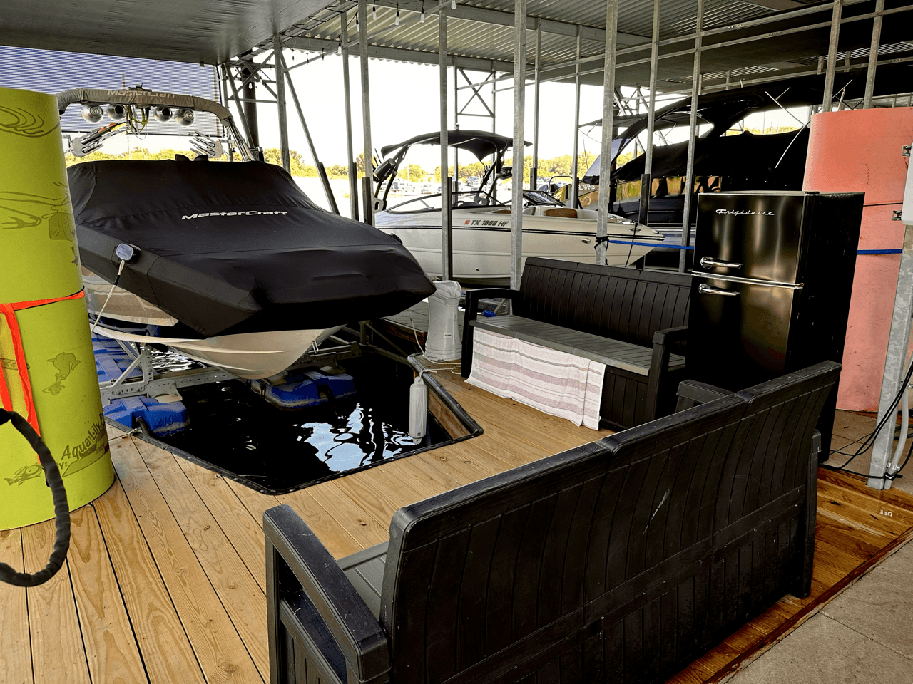 A covered boat dock features several moored boats, with some partially covered, alongside a seating area with black benches and a towel-draped cushion, all under a metal roof.