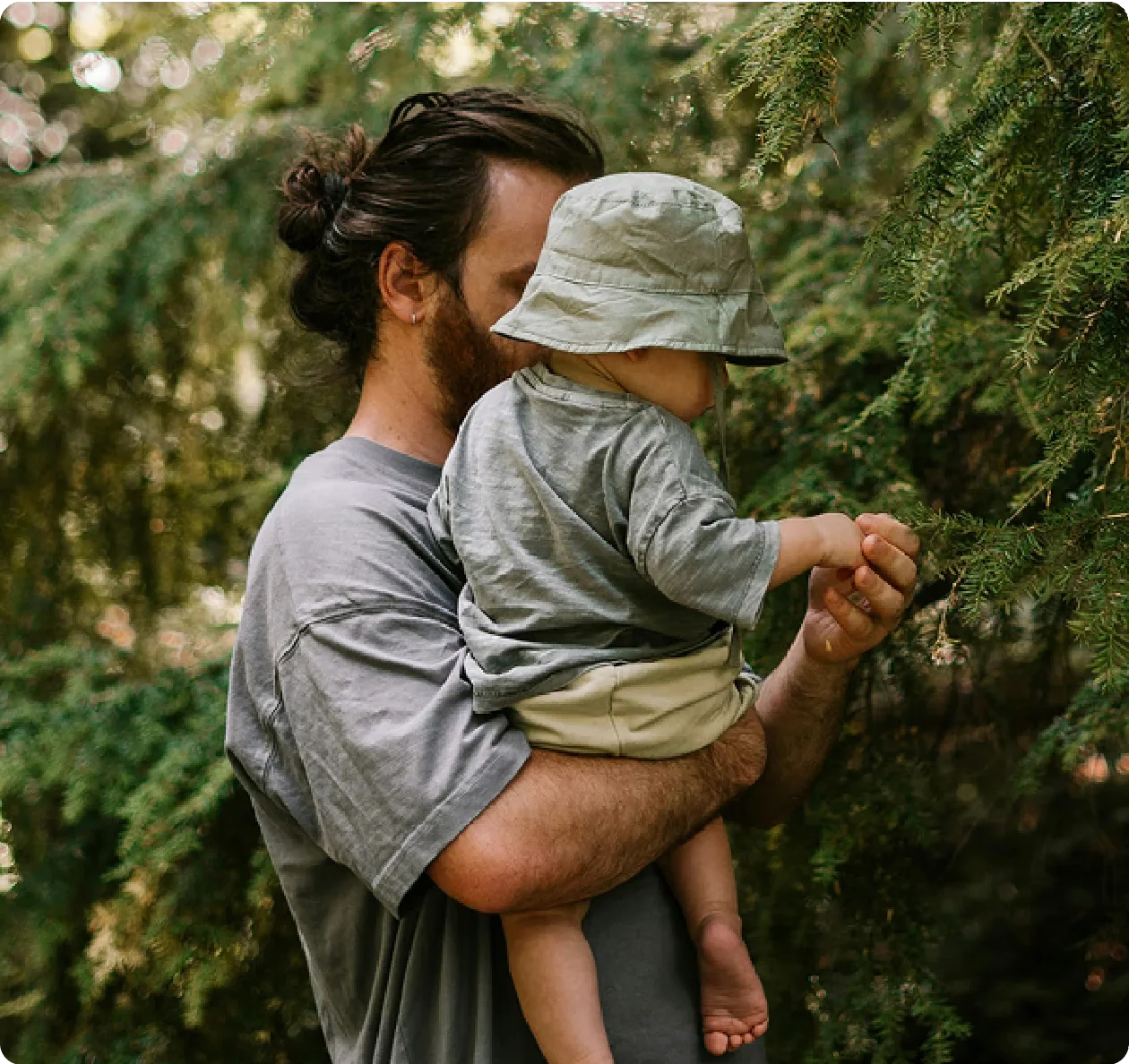 Man with a bun holds a child in a bucket hat near evergreen trees. The child reaches out to touch a branch.