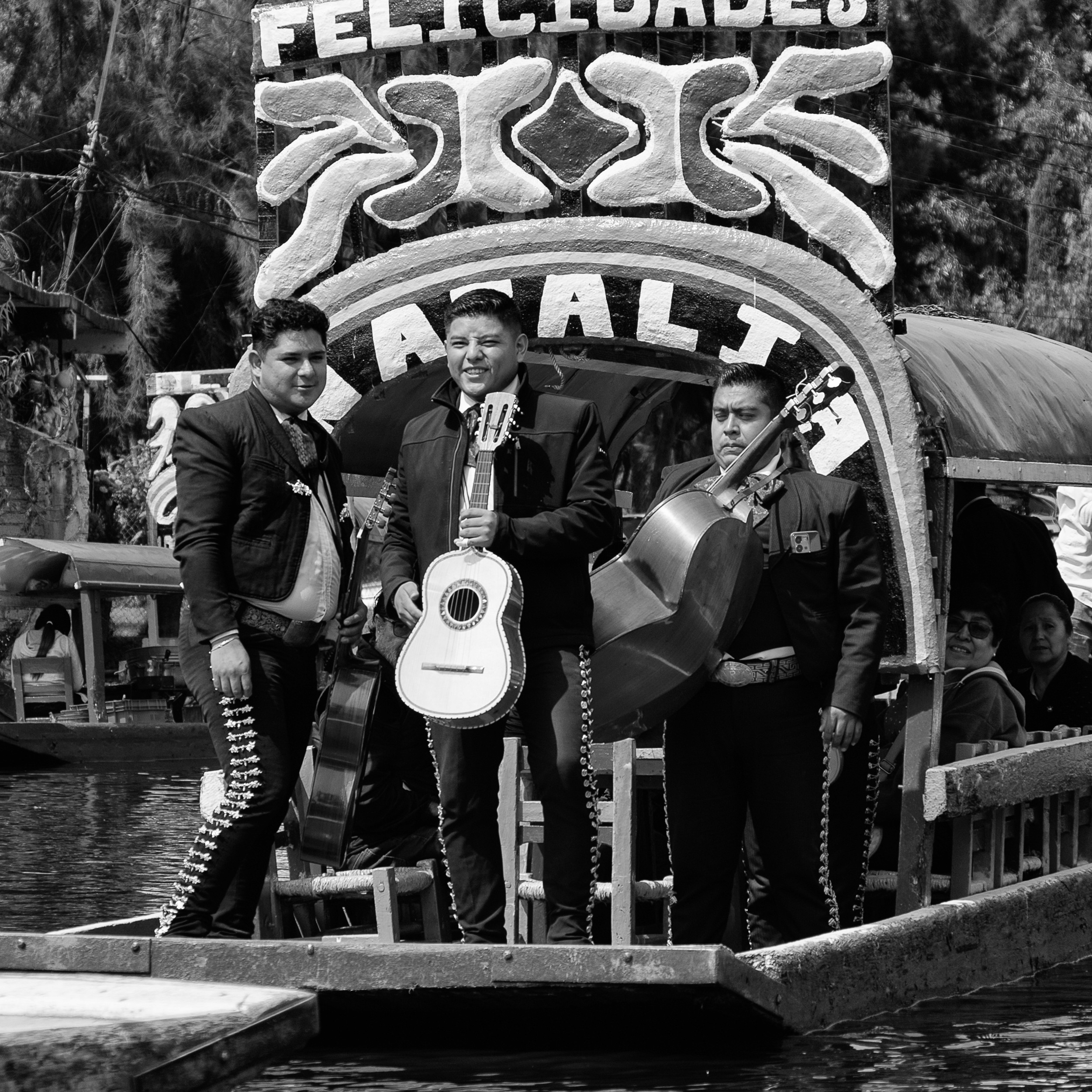 Black and white photograph of three mariachi musicians performing on a trajinera in Xochimilco, Mexico, under a sign that reads “Felicidades,” with passengers seated behind them on the boat.