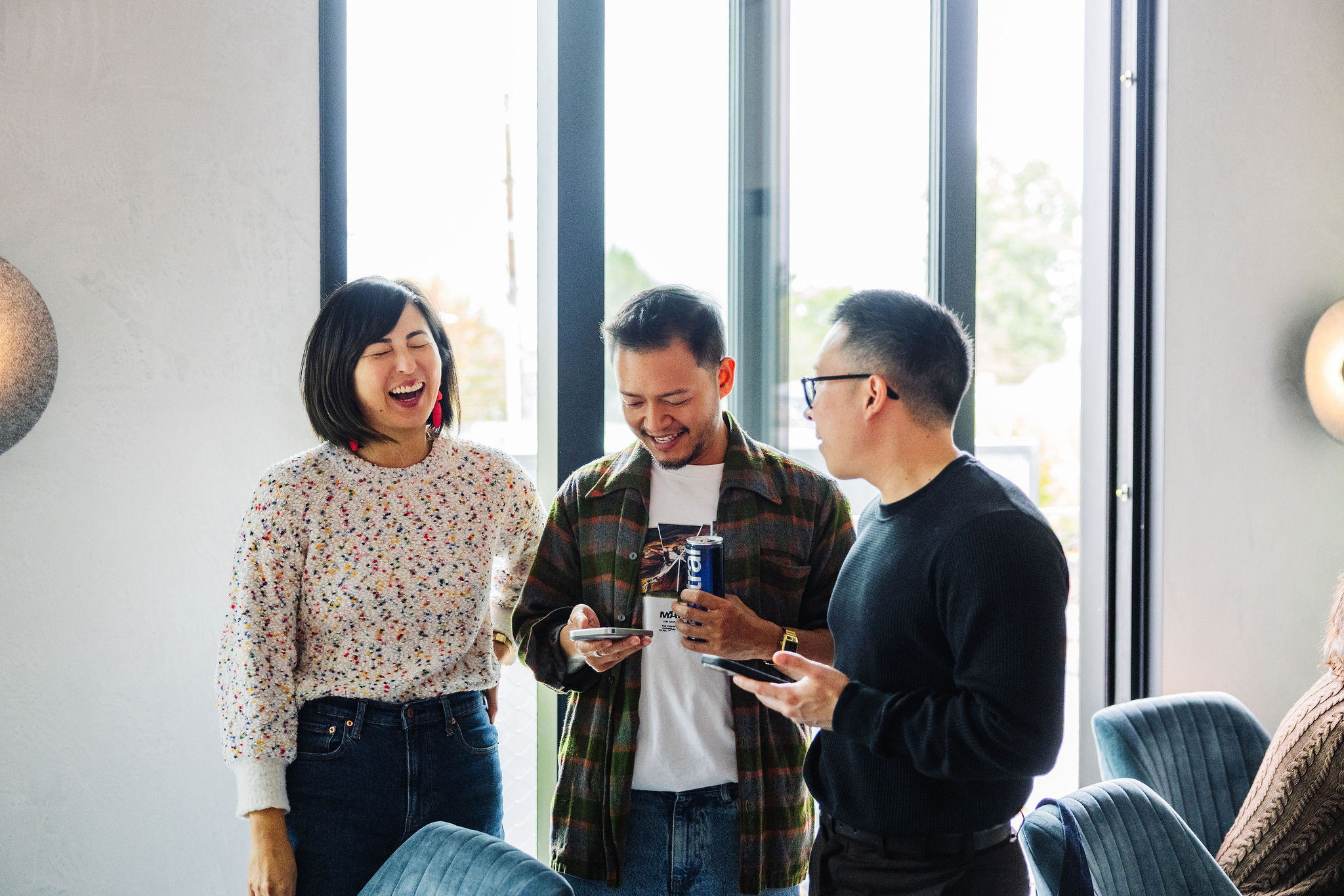 Three coworkers laughing together near large windows, looking at phones and holding a drink in a casual office setting.