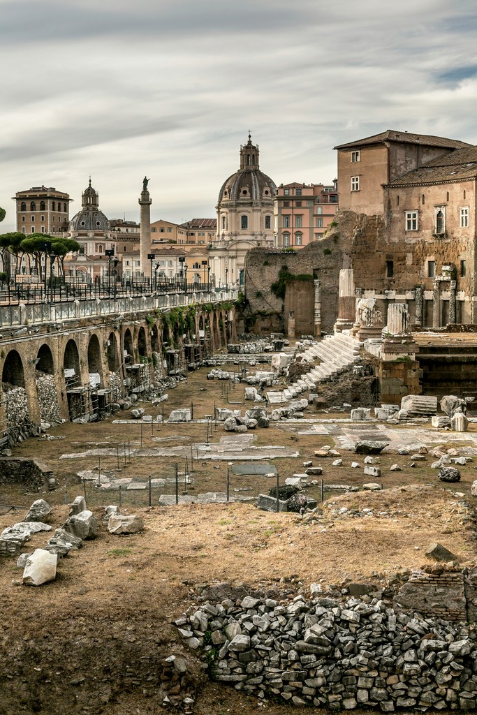 The Roman Forum, a historic site in Rome, Italy, showcases ancient ruins of monumental architecture, including columns, arches, and remnants of stone structures, with the cityscape and iconic domes of Renaissance-era buildings in the background under a cloudy sky.