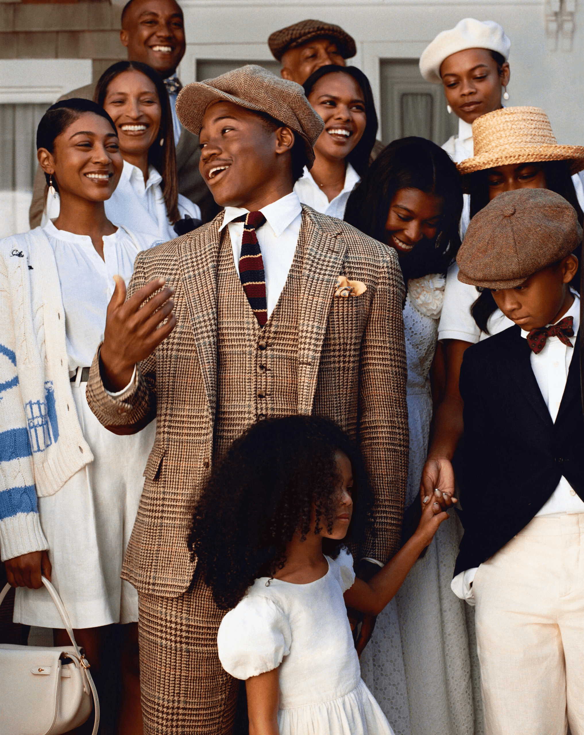 For Ralph Lauren’s Oak Bluffs Summer 2025 campaign, a well-dressed man in a brown plaid suit and tie is seen smiling among a group of adults and children in coordinated neutral outfits.
