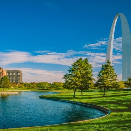 A large arch structure stands near a curved riverbank bordered by lush green grass and trees, with buildings in the background under a blue sky.