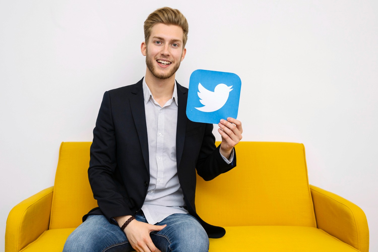  A man sits on a yellow couch, holding a Twitter sign, smiling as he engages with social media.