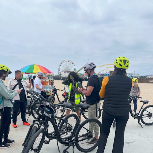 Un grupo de personas con cascos amarillos neón y bicicletas se reúne cerca de un vendedor con una colorida sombrilla junto a la playa, con una noria de fondo.