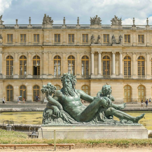 Eine Bronzestatue einer liegenden Figur mit einem Kind vor der prächtigen Fassade des Schlosses von Versailles.