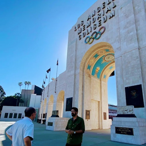 Two people stand outside the Los Angeles Memorial Coliseum, featuring Olympic rings and large archways, on a clear day.
