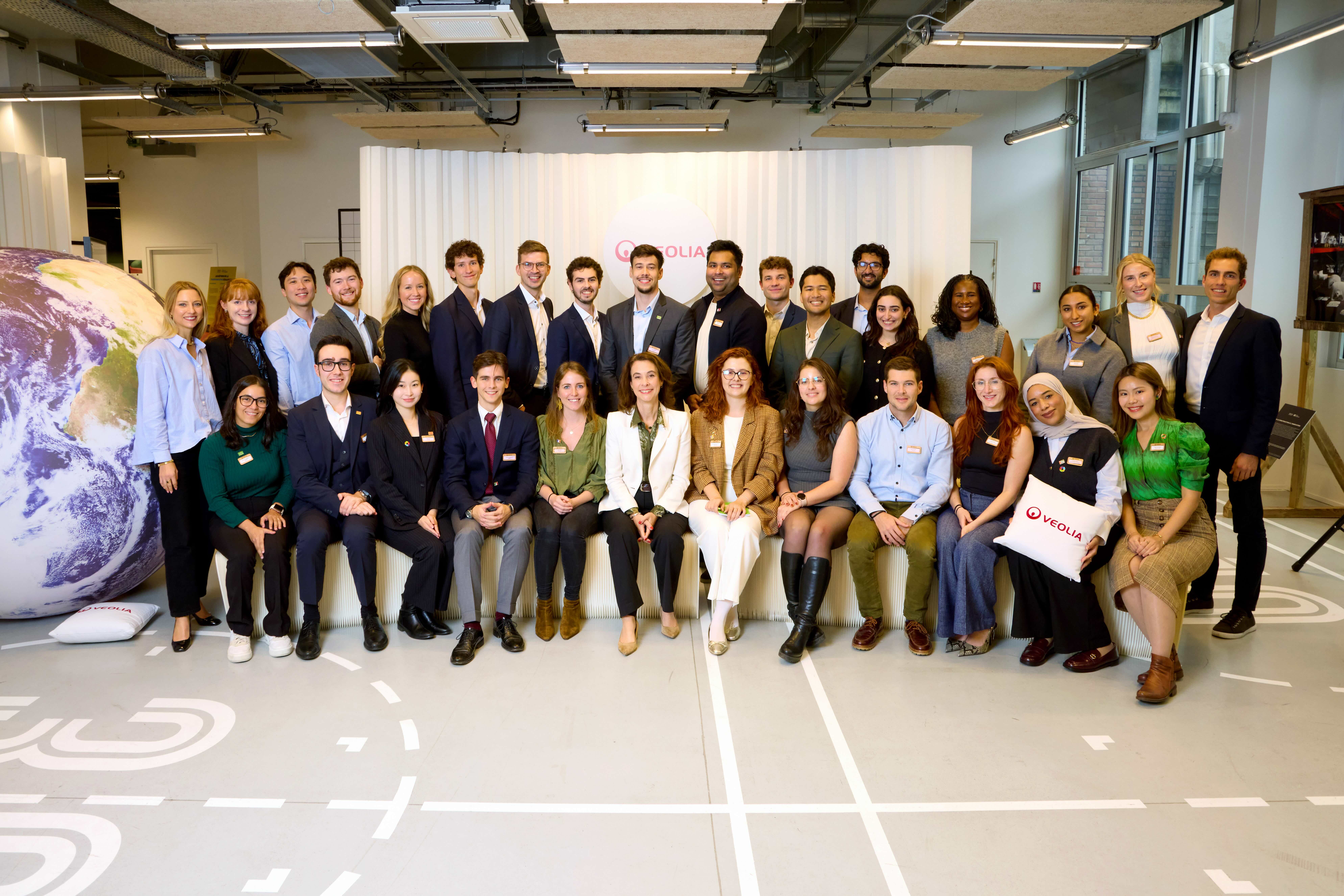 A young person with curly hair smiles at the camera while sitting in front of a laptop against a dark background.
