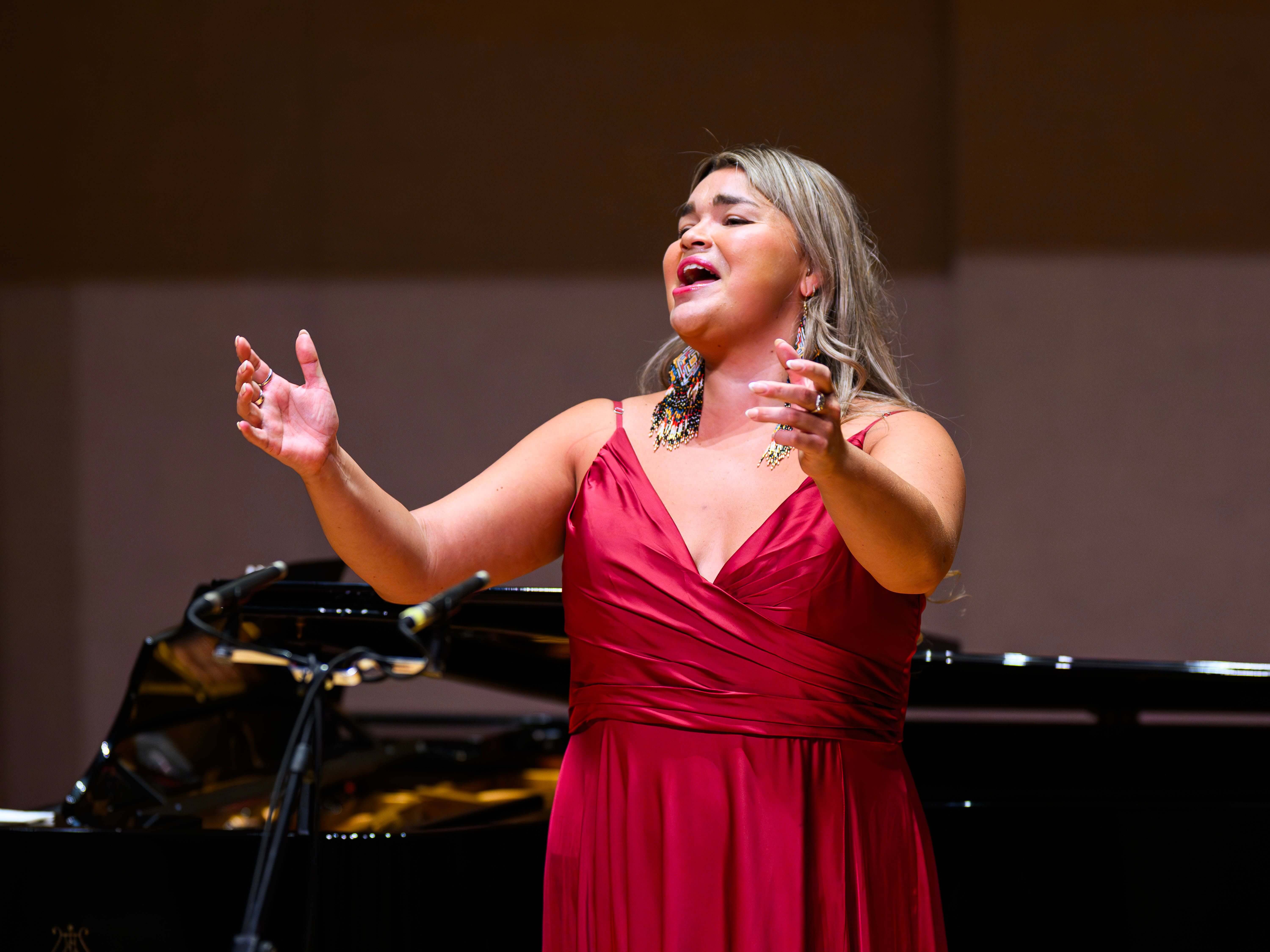 Elisabeth St-Gelais interprète une œuvre en récital, vêtue d’une robe rouge et de boucles d’oreilles perlées, devant un piano de concert.