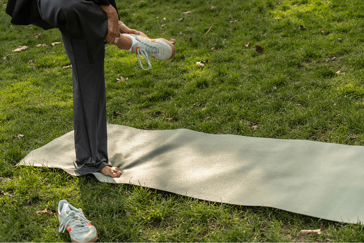A person stands on lush green grass, stretching one leg over a yoga mat while wearing casual sportswear, emphasizing a commitment to a healthy lifestyle.